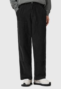 Pantaloni in corduroy neri con gambe ampie, dotati di grandi tasche anteriori e un motivo a coste testurizzato. Abbinati a sneaker di colore chiaro.