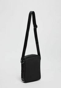 Borsa a tracolla nera con forma rettangolare, realizzata in tessuto liscio, dotata di chiusura con zip e tracolla regolabile. Design piatto, hardware minimale.