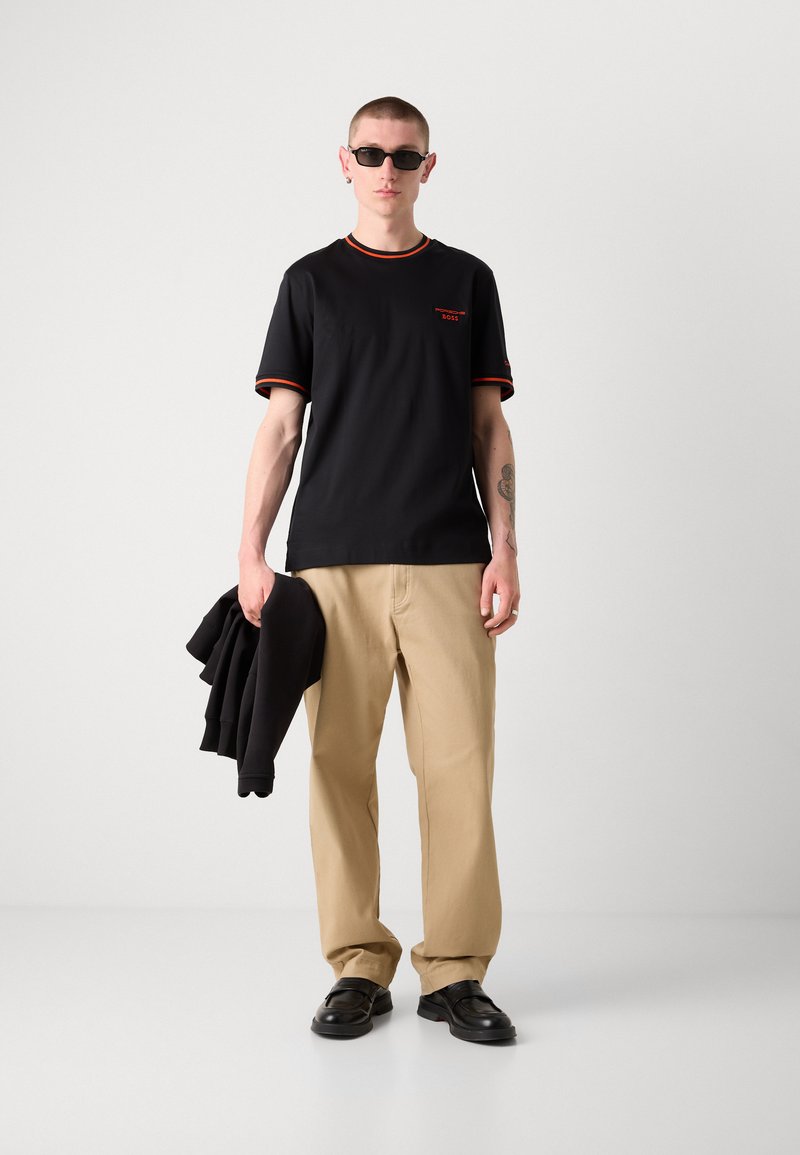 T-shirt noir à manches courtes avec une bordure rouge, associé à un pantalon beige. Le modèle tient un sweat-shirt noir, portant des lunettes de soleil et des chaussures noires.