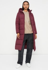 Geacă puffer bordo cu un design lung și glugă, cu închidere prin fermoar și model matlasat. Asortată cu un top bej și pantaloni cargo negri.