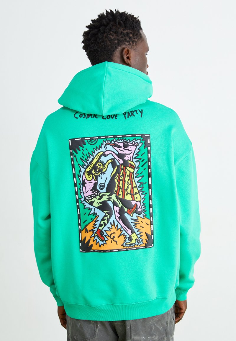 Grön hoodie med en grafisk illustration på baksidan, som visar livfulla färger och en dansscen. Texten ovanför lyder "COSMIC LOVE PARTY."