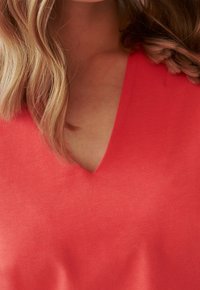 V-hals, kortærmet top i lys koralfarvet stof, glat tekstur, tætsiddende design, der fremhæver halsudskæring og skuldre.