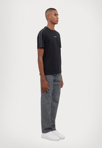 T-shirt noir à manches courtes avec un col rond, présentant des rayures blanches sur les côtés et un petit logo. Assorti à un jean droit gris et des baskets blanches.