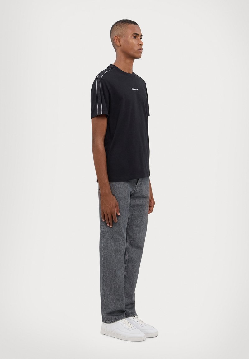 T-shirt noir à manches courtes avec un col rond, présentant des rayures blanches sur les côtés et un petit logo. Assorti à un jean droit gris et des baskets blanches.