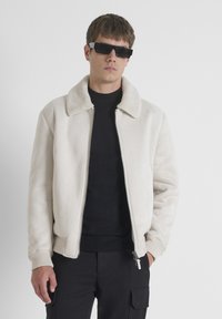 Svijetlosmeđa bomber jakna s mekim krznom na ovratniku, glatke teksture, sa zatvaračem sprijeda, rebrastim manžetama i džepovima. Nošena preko crnog džempera.