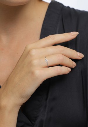 Silver ring with a central clear gemstone, delicate band, and minimalist design, worn on a hand with natural nails against a dark fabric background.