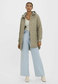 Veste longue matelassée vert olive avec capuche, dotée d'une fermeture à boutons, de poignets côtelés, associée à un jean bleu clair à jambes larges.