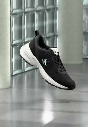 Baskets Calvin Klein noir et blanc avec logo sur le côté et la languette, flottant sur un fond industriel minimaliste avec des blocs de verre et un sol en béton.