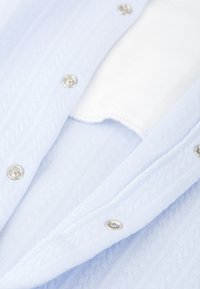 Camisa de punto azul claro con botones a presión plateados, que presenta un patrón texturizado y un forro de tela blanca en el cuello y las aberturas.