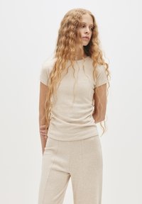 Beige toppi lyhythihaisena, valmistettu pehmeästä kankaasta, jossa on hienovaraiset rypytykset sivuilla ja pyöreä pääntie, yhdistettynä samansävyisiin housuihin.