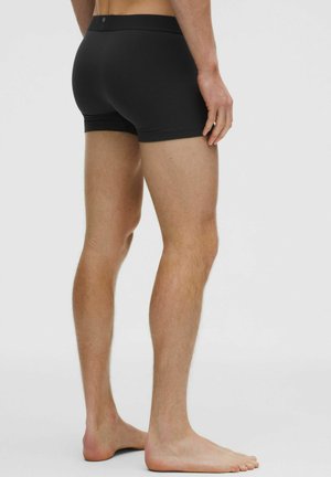 Czarne bawełniane bokserki o dopasowanym kroju, gładkiej fakturze i elastycznym pasku w talii. Model pokazany jest z boku, stojąc boso.
