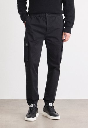 Mann trägt schwarze Cargo-Hosen mit elastischen Bündchen, seitlichen Klappentaschen, einen schwarzen Pullover und schwarze Sneaker mit weißen Sohlen.