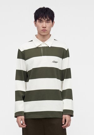 Joven con una camiseta de rugby de rayas anchas verdes y blancas de manga larga con cuello, de pie frente a un fondo liso.