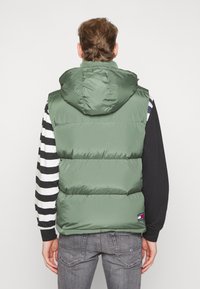 Zelená prošívaná vesta s kapucí, s hladkou texturou a horizontálním prošitím. Nositelná přes černobíle pruhované tričko s dlouhým rukávem.