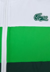 Vita, gröna och mörkgröna tygpaneler med en vit dragkedja och en grön krokodil-logotyp på övre högra sidan.