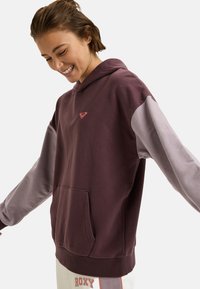 Joven sonriendo, vistiendo una sudadera color burdeos y gris claro con un pequeño logotipo de corazón y pantalones blancos con el texto "ROXY".