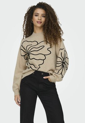 Beige trui met zwarte bloemenborduursel, geribbelde mouwen en een ronde hals. Gecombineerd met hooggetailleerde zwarte jeans, die een ontspannen pasvorm laten zien.