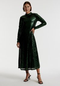 Groene pailletten jurk met lange mouwen en hoge halslijn, met een vloeiende silhouet en een zoom net boven de enkels. Draag met zwarte sandalen met straps.