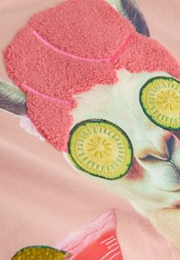 Visage de chat avec des tranches de concombre vertes sur les yeux et un tissu rose texturé ressemblant à un couvre-chef sur un fond rose clair.