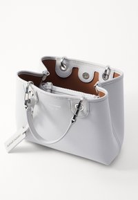 Biała skórzana torebka o fakturowanej powierzchni, srebrnych okuciach, podwójnych uchwytach oraz brązowym wnętrzu. Posiada zapięcie na zamek błyskawiczny oraz detal z logo.