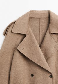 Beige wool coat with wide lapels, double-breasted buttons, and shoulder epaulette detail on plain background.