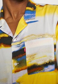 Col et poche poitrine d'une chemise colorée avec des motifs abstraits jaunes, bleus, marron et noirs et une petite étiquette rouge Levi's sur la poche.