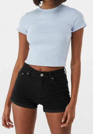 Kvinde iført lyseblå kortærmet croppet top og sorte højtaljede denimshorts med opbukket kant, stående mod en ensfarvet baggrund.