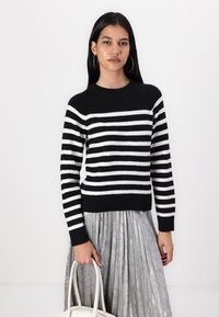 Mustat ja valkoiset raidalliset neuleet, joissa on pyöreä pääntie ja ribbikuvat, yhdistettynä litsittyyn hopeahameeseen ja valkoiseen käsilaukkuun.