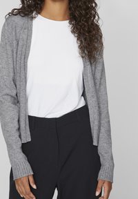 Ljusgrå cardigan med ribbade manschetter, bärs över en vit t-shirt. I kombination med mörka byxor vilket ger en enkel och avslappnad stil.