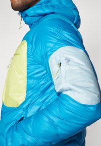 Plava puffer jakna s svijetložutom džepom, koja ima kapuljaču, reljefnu tkaninu i džep na rukavu s patentnim zatvaračem, ukrašen zelenim akcentima.