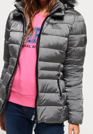 Mujer con chaqueta acolchada gris brillante con capucha con ribete de pelo sobre una sudadera rosa brillante y pantalones vaqueros azules.