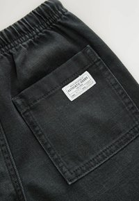Sorte stofbukser med elastisk talje og baglomme. Lommen har en hvid etikette med teksten "AUTHENTIC GOODS". Struktureret overflade.