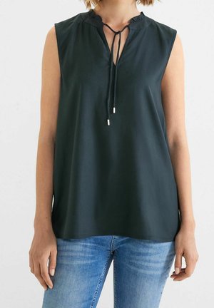 Vrouw draagt een mouwloze zwarte blouse met een striksluiting bij de hals en een blauwe spijkerbroek, staand tegen een effen lichte achtergrond.