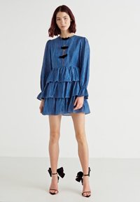 Vestido azul riscado em camadas com mangas longas, decote com fenda e botões pretos. Combinado com sandálias de salto alto pretas com detalhes de laço.