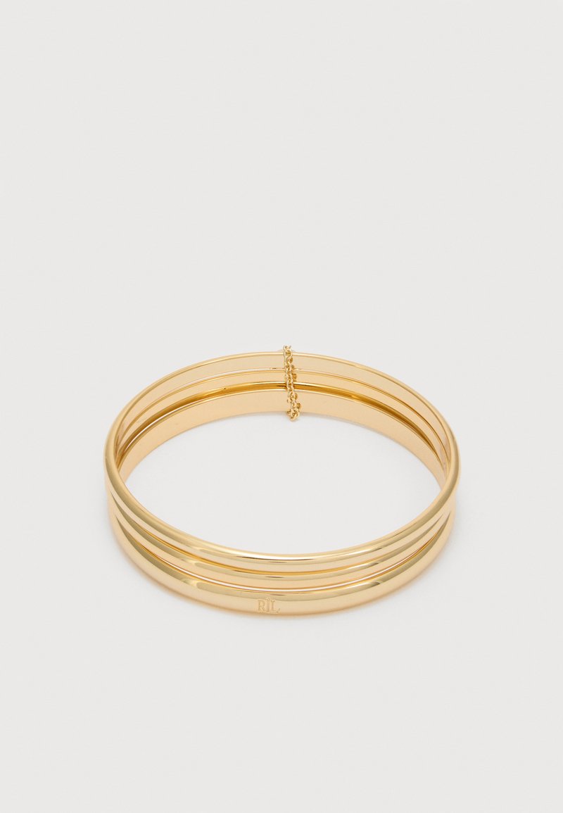 Βραχιόλι με τριπλή βέργα σε χρυσό τόνο, με μικρή αλυσίδα ασφαλείας και λείο γυαλιστερό φινίρισμα, τοποθετημένο επίπεδα σε ουδέτερο φόντο.