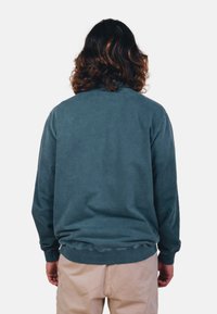 Felpa in teal scuro con vestibilità rilassata, polsini e orlo a coste, e un tessuto testurizzato. Presenta un colletto classico ed è abbinata a pantaloni chiari.