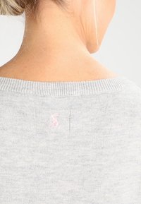 Gros plan sur le cou et le haut du dos d'une femme blonde portant un pull gris clair avec un petit logo rose brodé.