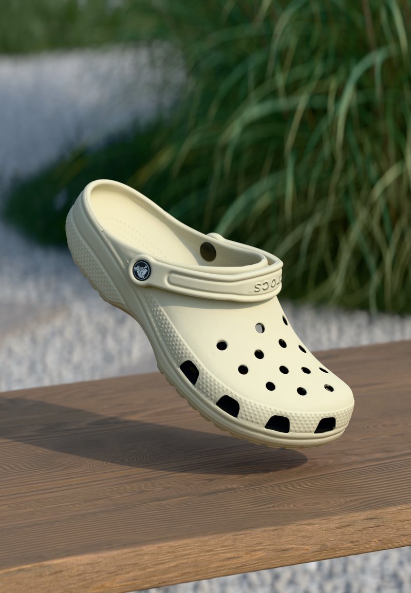 Sabot beige à enfiler avec des trous de ventilation et une bride au talon, posé sur une surface en bois, environnement extérieur avec des plantes vertes en arrière-plan.