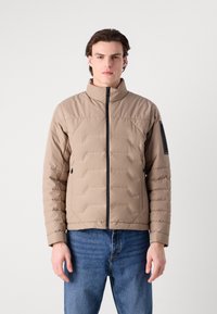 Beige pufferjacka med horisontella sömmar, hög krage och främre dragkedja. Har fickor med dragkedja och kontrasterande svarta detaljer på ärmen.