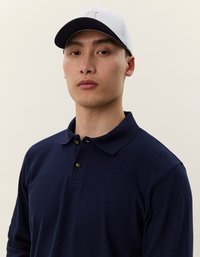 Tumši zils garām piedurknēm polo krekls ar apkakli un trim pogu rindām; apvienots ar baltu un tumši zilu beisbola cepuri ar logotipu.