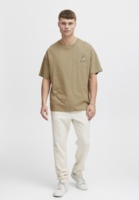 Khaki bomulds t-shirt med løs pasform, der har et lille broderet design. Kombineret med hvide tapered bukser og hvide sneakers.