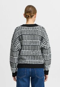 Raksts ar tekstūru melnbaltā džemperī ar ģeometrisku rakstu, apaļu kakla izgriezumu un ribotu piedurkņu un apakšmalu. Aizmugures skats rāda brīvu piegriezumu.