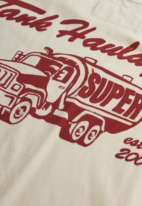 Camiseta beige con un gráfico rojo de un camión cisterna que dice 'Super' y 'Tank Haulers est. 2000'. Líneas audaces y diseño simple.
