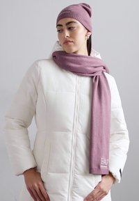 Berretto di lana rosa e sciarpa coordinata indossati sopra un giubbotto bianco imbottito. La sciarpa presenta dettagli con il logo. Il tessuto sembra morbido e caldo.