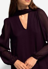 Diep paarse blouse met een V-hals, lange doorzichtige mouwen en gathered manchet. Heeft een zachte, gestructureerde stof en een brede halsdoek.