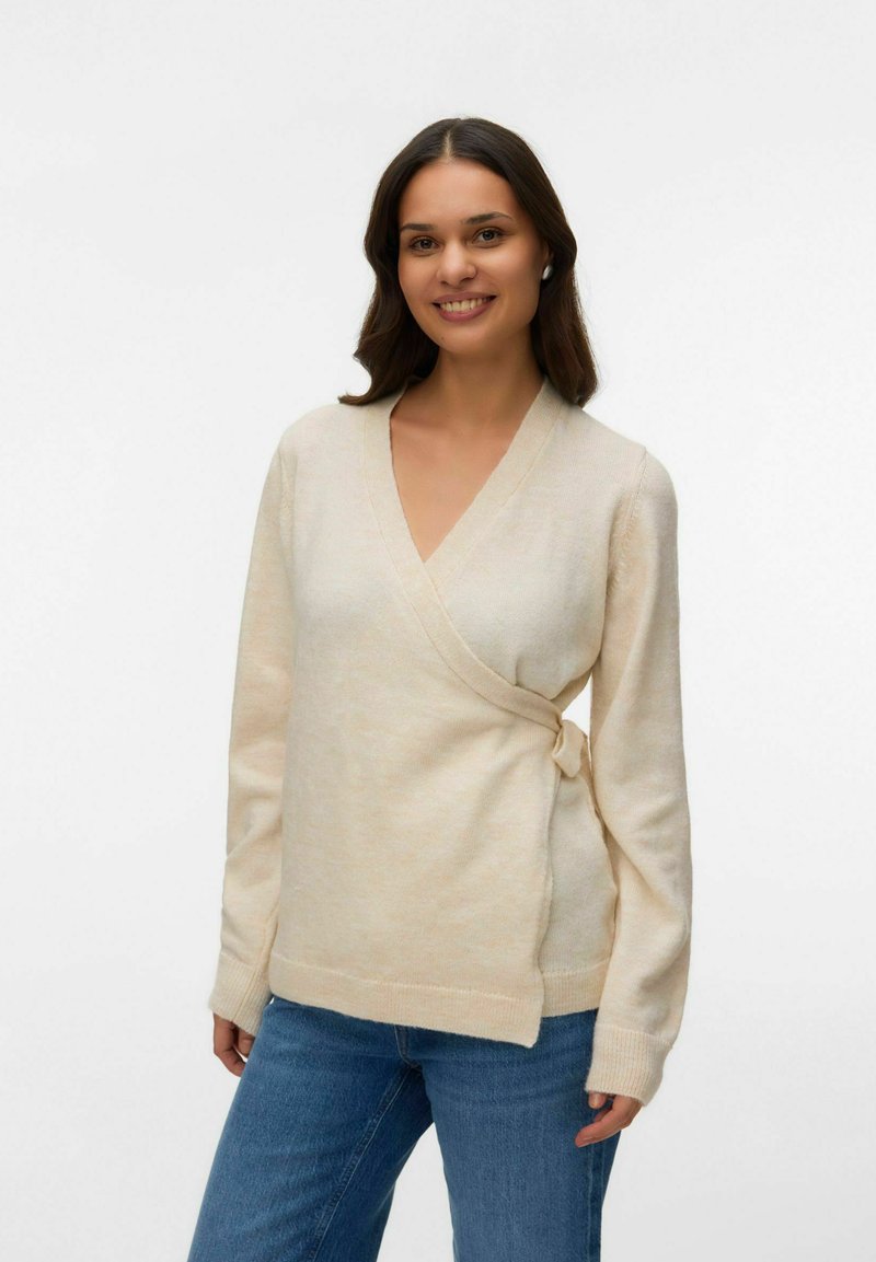 Junge Frau lächelnd, trägt einen cremefarbenen Wickelpullover mit langen Ärmeln und blaue Jeans, steht vor einem schlichten Hintergrund.