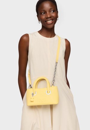 Mujer sonriente con vestido beige sin mangas y manos en los bolsillos, lleva un pequeño bolso amarillo con cadena cruzado sobre el cuerpo.