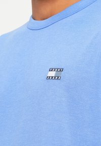 Világoskék pamut póló kerek nyakkivágással, amelyen egy téglalap alakú hímzett logó található "TOMMY JEANS" felirattal a mellkasán.