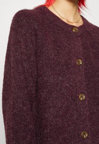 Vinröd stickad cardigan med rund hals, texturerad tyg och fyra stora bruna knappar fram.