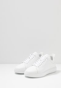 Sneakers bianchi in pelle con punte arrotondate, lacci piatti e alette di sospensione; suole in gomma testurizzata; design minimalista con dettagli di cucitura.